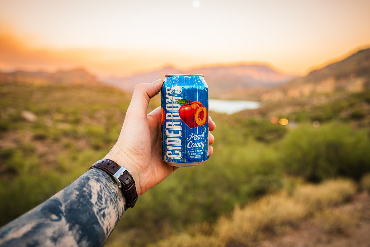 Hand holding a blue Ciderboys Peach County can with hills and a lake softly blurred in warm evening light