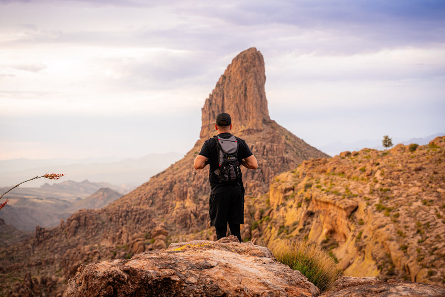 A hiker wearing a black backpack looking toward Weavers Needle beneath a cloudy sky