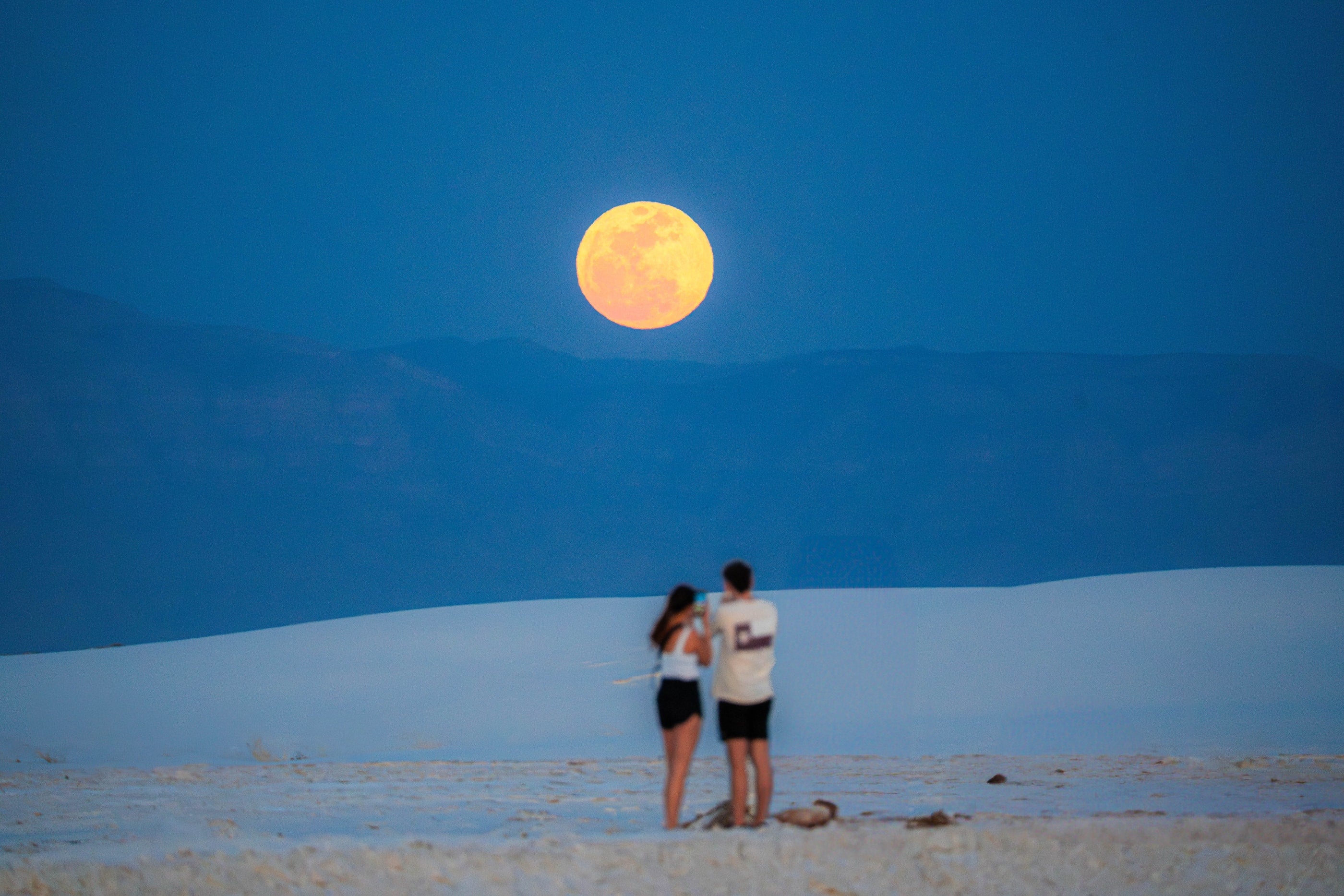 A full moon rising above blue mountains and white dunes with visitors below